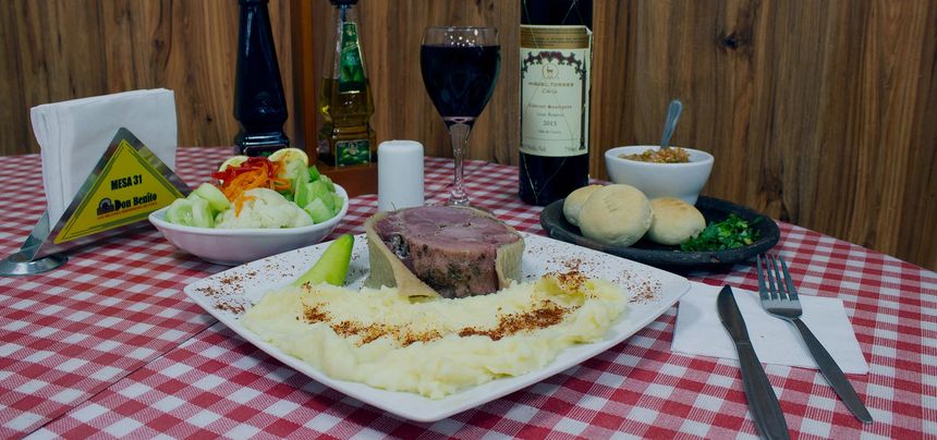 Mesa servida con arrollado huaso, puré, ensalada, pan amasado y una copa de vino sobre el mantel a cuadros.